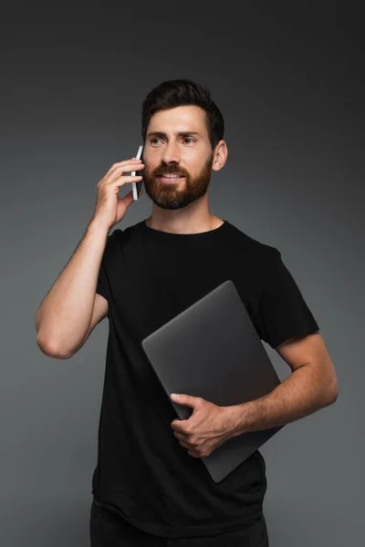 Счастливый бородатый мужчина в черной футболке держа ноутбук, разговаривая на смартфоне изолированы на сером — стоковое фото