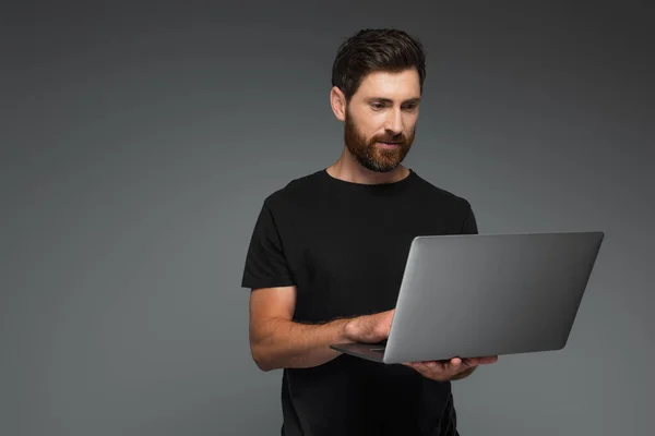 Bearded man in black t-shirt using laptop isolated on grey — Foto stock