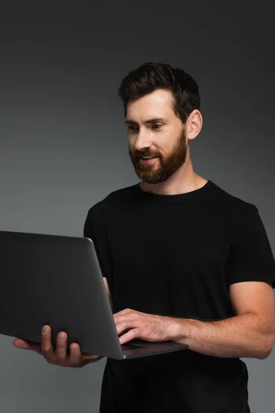 Cheerful and bearded man in black t-shirt using laptop isolated on grey — Photo de stock