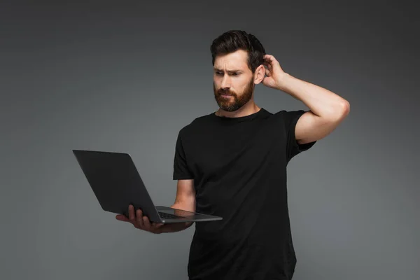 Confused freelancer in black t-shirt using laptop isolated on grey — Stock Photo