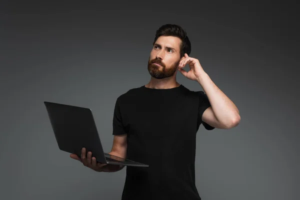 Pensive freelancer with beard holding laptop and looking away isolated on grey - foto de stock