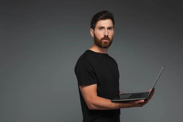 Бородатий фрілансер у чорній футболці тримає ноутбук ізольовано на сірому — стокове фото