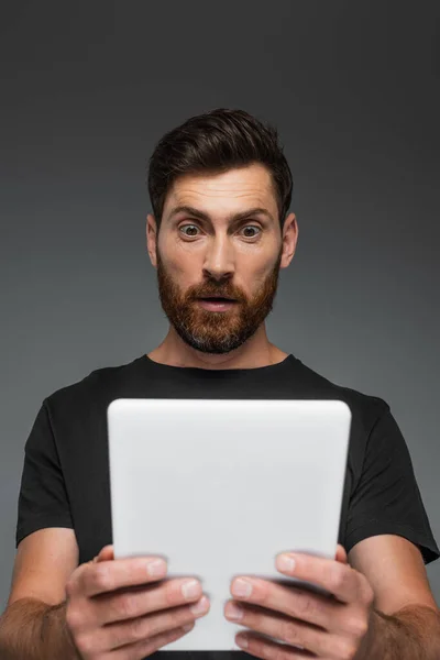 Shocked and bearded man in black t-shirt holding digital tablet isolated on grey — Stockfoto