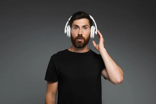 Surprised man touching wireless headphones and listening music isolated on grey — Stock Photo