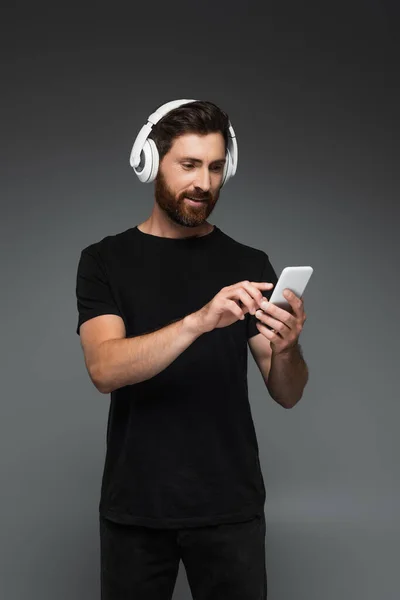 Cheerful man in wireless headphones listening music and using smartphone isolated on grey — Stockfoto