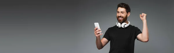 Возбужденный человек в беспроводных наушниках с помощью смартфона изолированы на серый, баннер — стоковое фото