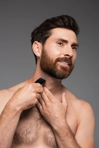 Portrait of bearded and shirtless man with hair on chest shaving with electric razor isolated on grey — Photo de stock