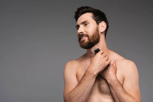 Cheerful and shirtless man with hair on chest shaving with electric razor isolated on grey — Stockfoto