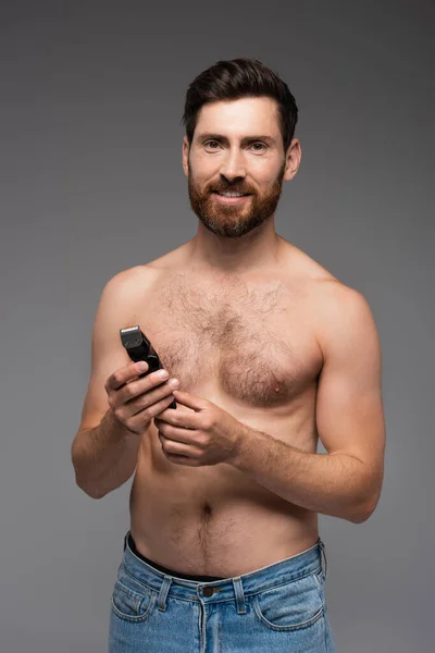 Щасливий і без сорочки чоловік з волоссям на грудях тримає електричну бритву ізольовано на сірому — стокове фото