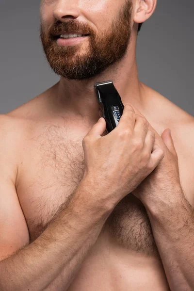 Cropped view of shirtless man smiling and shaving beard with electric razor isolated on grey — Foto stock