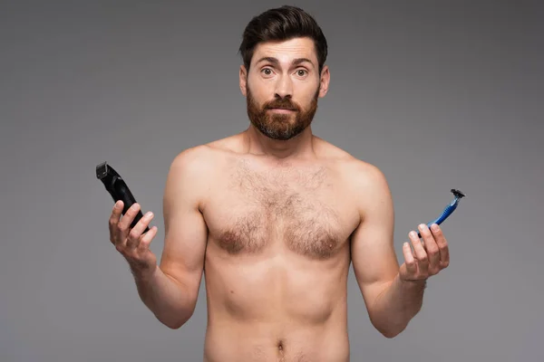 Збентежений і без сорочки чоловік з бородою тримає безпеку і електричні бритви ізольовані на сірому — стокове фото