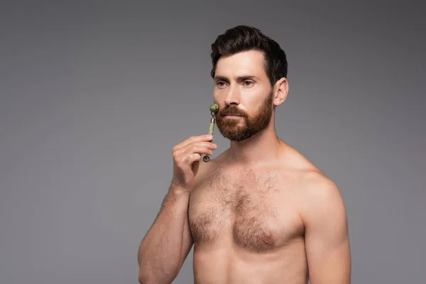 Shirtless man with beard using jade roller while massaging cheek isolated on grey — Foto stock