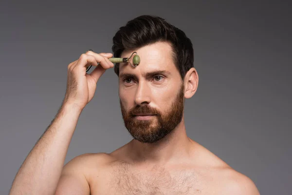 Shirtless man with beard using jade roller while massaging forehead isolated on grey - foto de stock