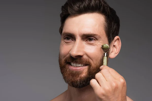 Happy man with beard using jade roller while massaging face isolated on grey — Stock Photo