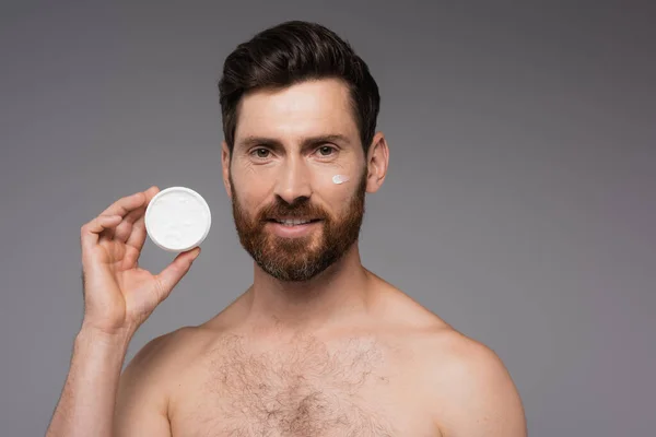 Bearded man with cream on face smiling while holding container isolated on grey — Stock Photo