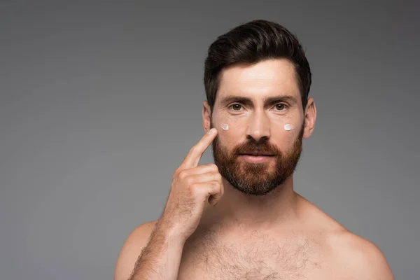 Bearded man pointing with finger at cream on face isolated on grey — Stock Photo