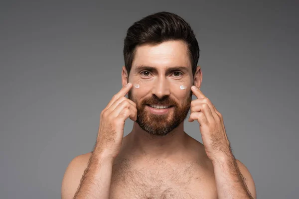 Бородатий чоловік наносить крем на обличчя і посміхається ізольовано на сірому — стокове фото