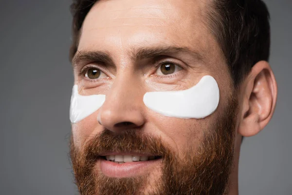 Крупный план жизнерадостного бородатого мужчины с увлажняющими глазными заплатами, изолированными на сером — стоковое фото