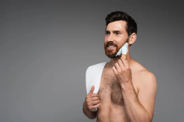 Happy bearded man with towel applying shaving foam with shaving brush isolated on grey — Photo de stock