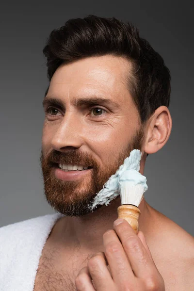 Портрет щасливого бородатого чоловіка, який наносить піну для гоління з пензлем для гоління ізольовано на сірому — стокове фото