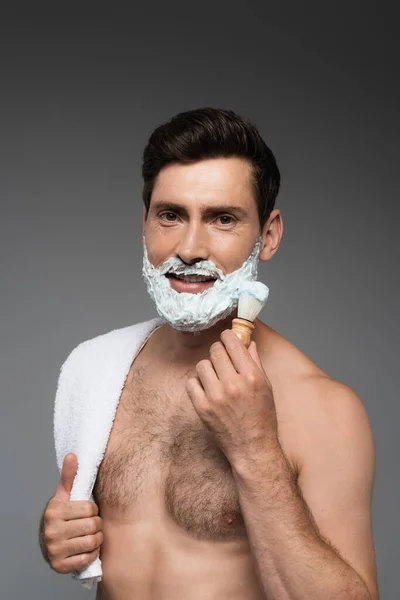 Веселий чоловік наносить білу піну для гоління на обличчя і посміхається ізольовано на сірому — стокове фото