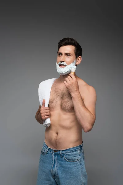 Shirtless man in jeans applying white shaving foam on face on grey — Stockfoto