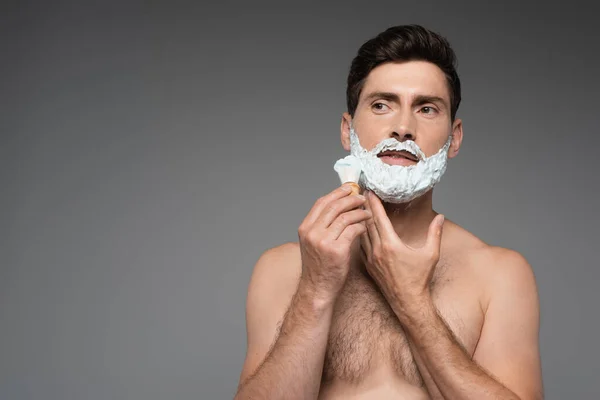 Shirtless man applying white shaving foam on face on grey — Stockfoto