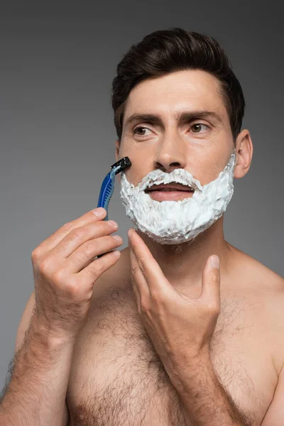 Shirtless man with white shaving foam on face shaving with safety razor on grey — Stockfoto
