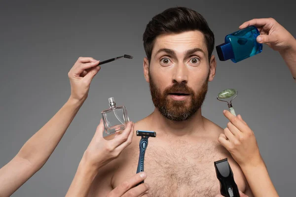 Women holding beauty products around bearded and surprised man on grey — Photo de stock