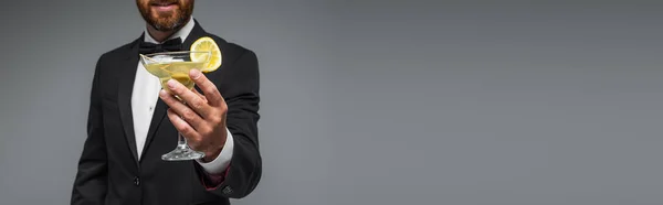 Обрізаний вид веселого і бородатого чоловіка в костюмі з краваткою з бантом, що тримає склянку коктейлю ізольовано на сірому, банер — стокове фото