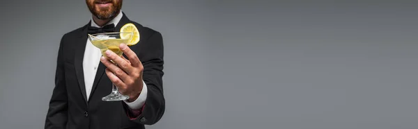 Cropped view of cheerful and bearded man in suit with bow tie holding glass of cocktail isolated on grey, banner — Foto stock