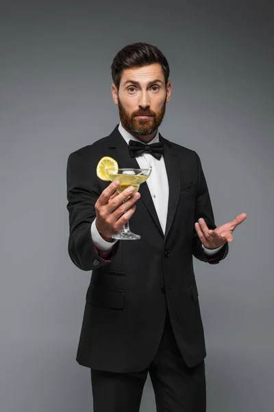 Bearded man in elegant suit with holding glass with cocktail and gesturing isolated on grey — Fotografia de Stock