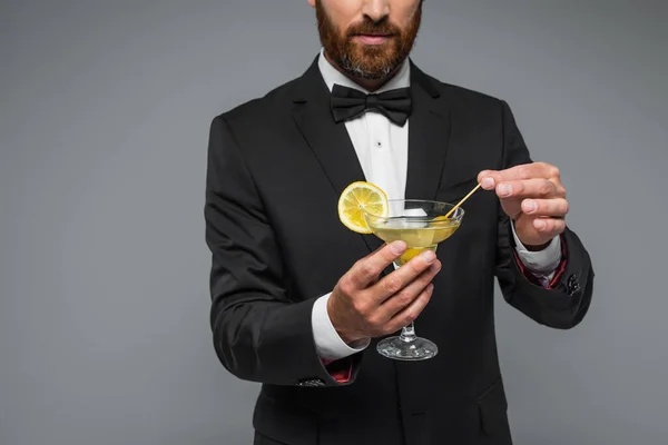 Partial view of bearded man in suit with bow tie holding toothpick with olive in glass with martini isolated on grey — Stockfoto