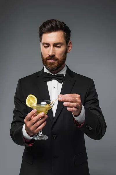 Bearded man in suit with bow tie holding toothpick with olive in glass with martini isolated on grey — Fotografia de Stock