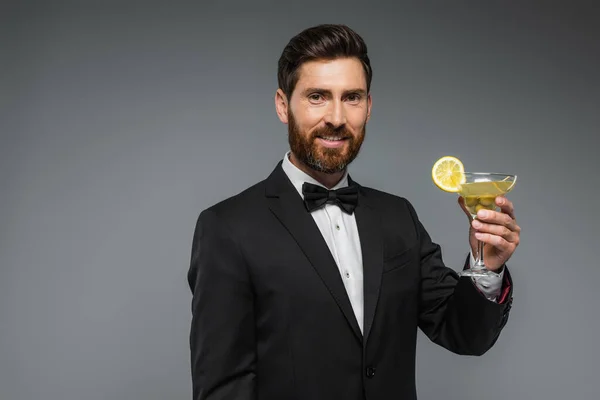 Щасливий чоловік в елегантному костюмі з краваткою, що тримає склянку коктейлю ізольовано на сірому — стокове фото
