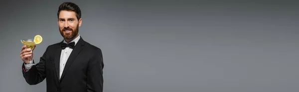 Cheerful man in elegant suit with bow tie holding glass with cocktail isolated on grey, banner — Photo de stock