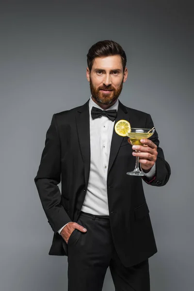 Bearded man in elegant suit holding glass with cocktail and posing with hand in pocket isolated on grey — Stock Photo