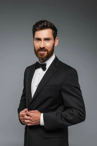 Щасливий і успішний чоловік з бородою, що стоїть в елегантному костюмі з краваткою ізольовано на сірому — стокове фото