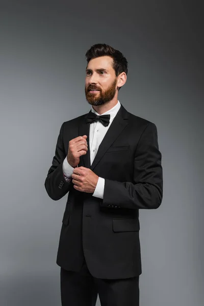 Улыбающийся мужчина с бородой, стоящий в элегантном костюме с галстуком-бабочкой, изолированным на сером — стоковое фото