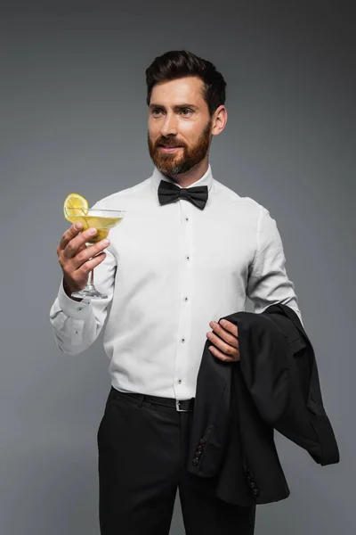 Bearded man in tuxedo with bow tie holding glass with cocktail and blazer isolated on grey — Stock Photo