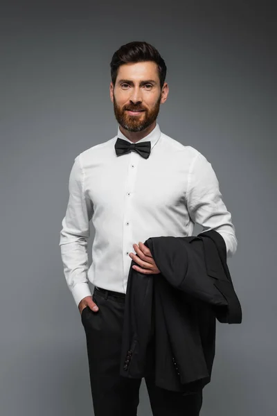 Cheerful man in white shirt with bow tie holding blazer and standing with hand in pocket isolated on grey — Stockfoto