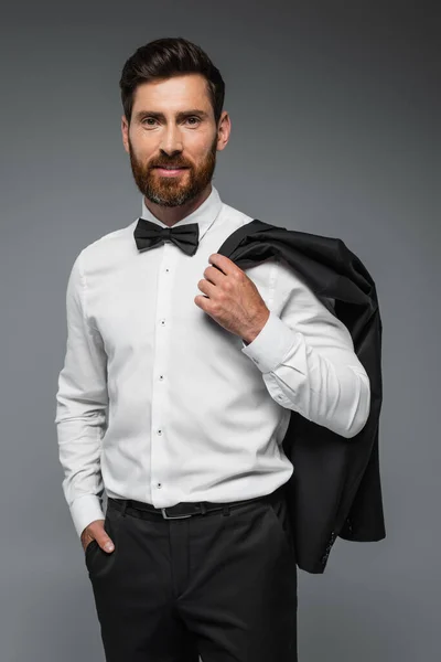 Cheerful man in suit with bow tie holding blazer and standing with hand in pocket isolated on grey — Fotografia de Stock