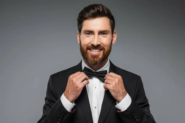 Bearded man in suit smiling and adjusting bow tie isolated on grey — Foto stock