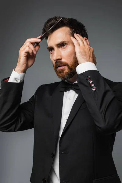 Bearded man in elegant tuxedo brushing hair with comb isolated on grey — Foto stock