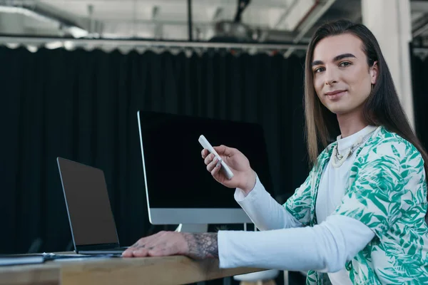 Queer designer using devices and smiling at camera in office — Stock Photo