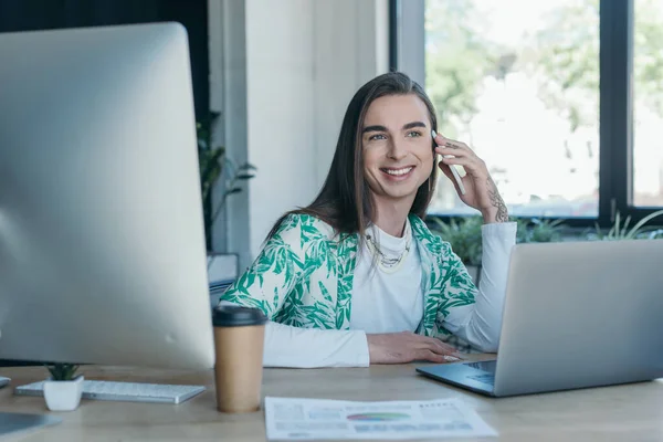 Cheerful queer designer talking on smartphone near devices and coffee in office - foto de stock