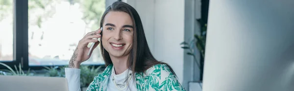 Smiling nonbinary designer talking on smartphone near devices in office, banner - foto de stock