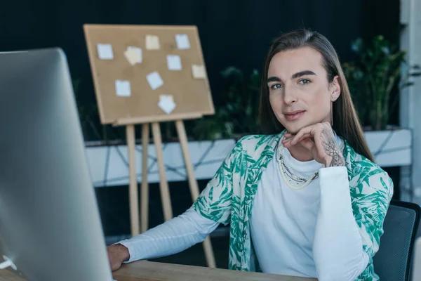 Tattooed queer designer looking at camera near computer in creative agency - foto de stock