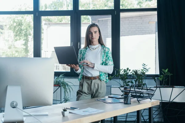 Queer designer holding laptop and looking at camera in creative agency — Stock Photo