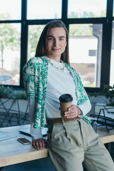 Smiling queer designer holding paper cup near working table in creative agency — Foto stock